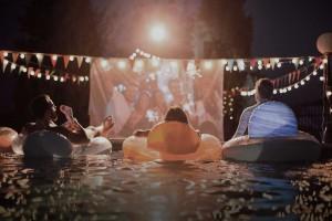 Adults lounging in inner tubes in pool at night screening a movie.